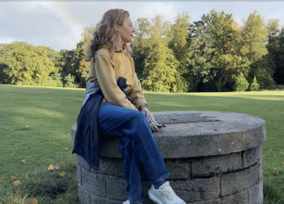 A picture of Clare Johnston seated on a stone well with a rainbow behind her