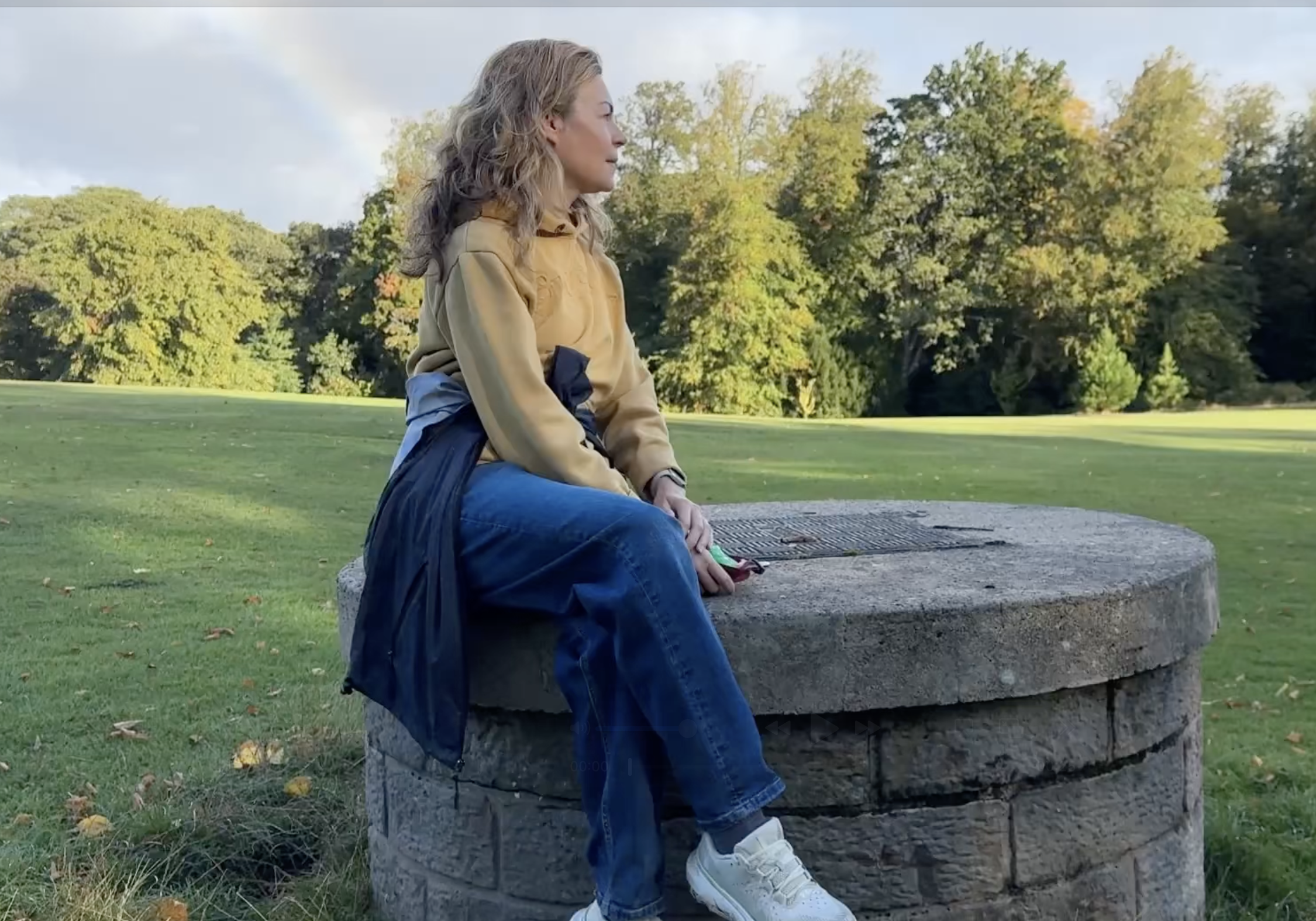 A picture of Clare Johnston seated on a stone well with a rainbow behind her