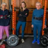Rhoda Stephen, Clare Johnston and Michael Stephen standing by their barbell training rack